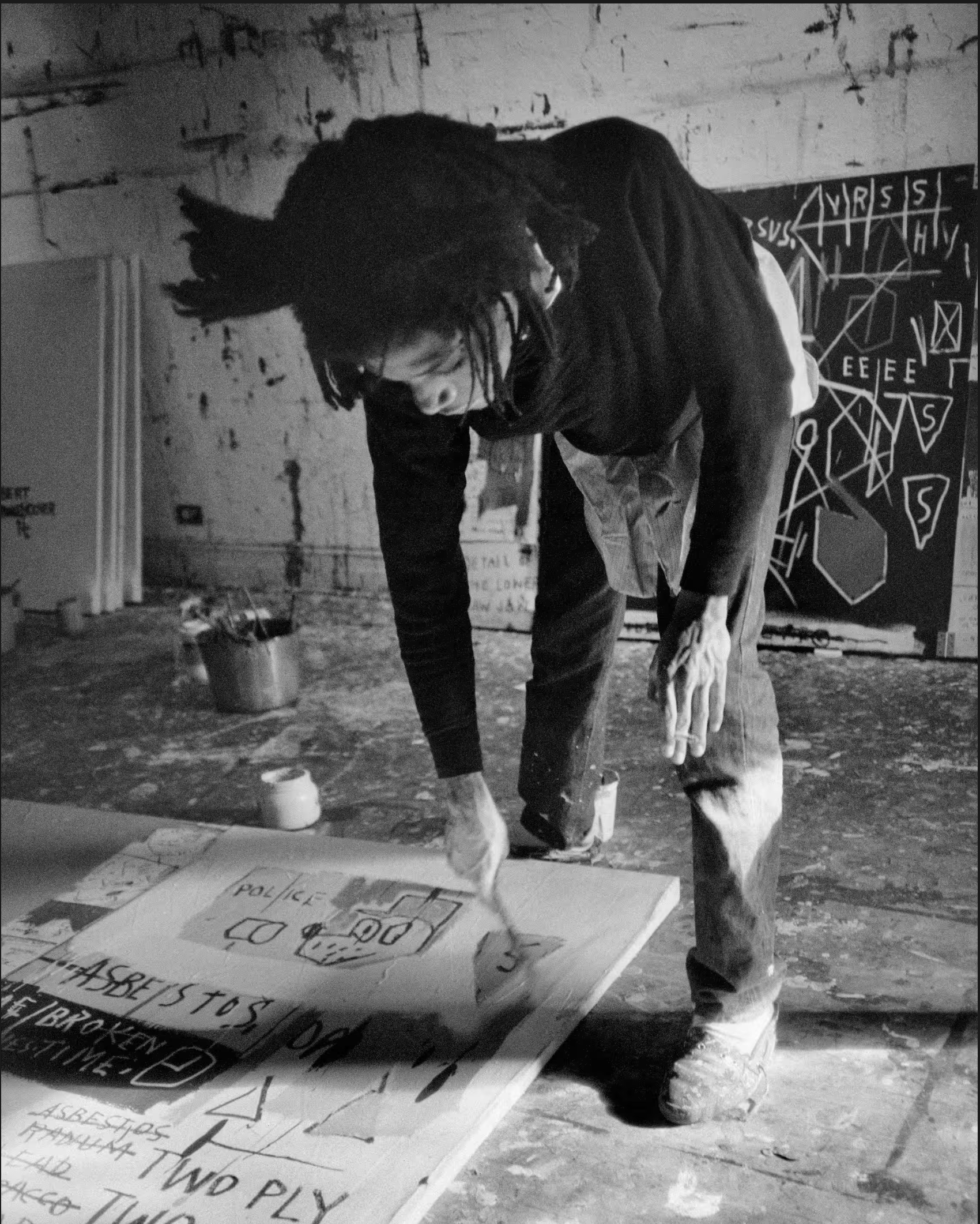 A man is bent down with a paint brush in hand painting on a canvas in a studio.